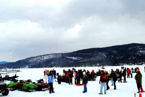 A helicopter hovers above a crowd on a snowy lake, with snowmobiles parked nearby and mountains in the background.