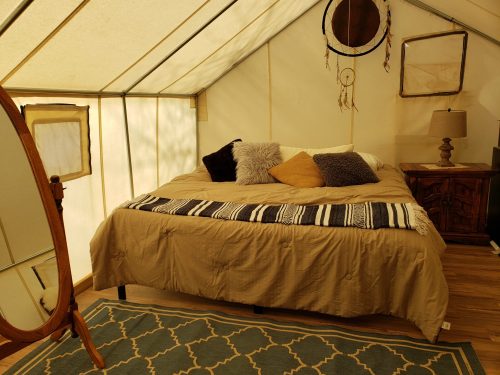 Cozy tent interior with a bed, decorative pillows, a lamp, and a mirror, featuring warm, earthy tones.
