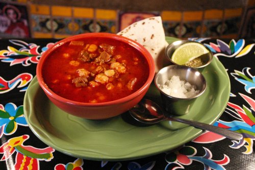 A bowl of hearty soup with meat and corn, served with tortillas, lime, onions, and spices on a colorful table.