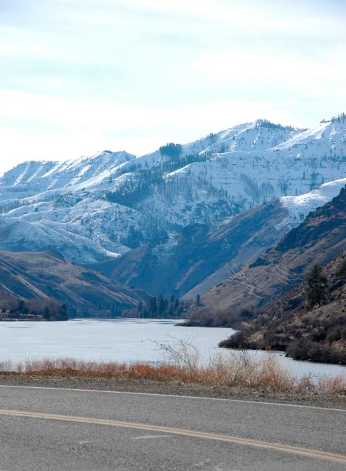 A scenic view of snow-capped mountains beside a calm river, with a road in the foreground.