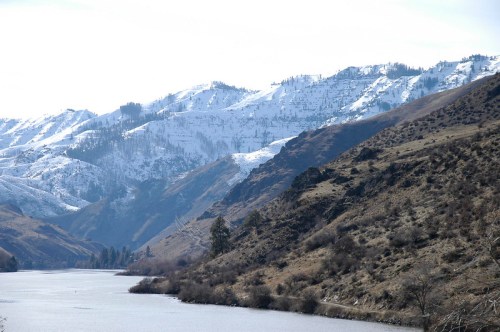 Snow-capped mountains rise above a calm river, surrounded by rugged terrain and sparse vegetation.