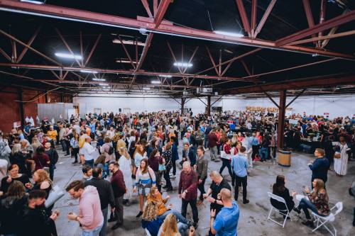 A large crowd of people mingles in a spacious industrial venue, with tables and chairs set up around the area.