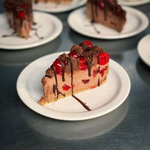 A slice of chocolate cherry cheesecake on a white plate, drizzled with chocolate and topped with cherries.