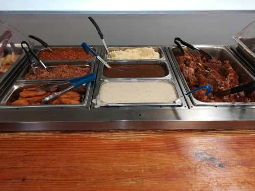 A buffet spread featuring various dishes, including pulled pork, mashed potatoes, gravy, and fried items, with serving tongs.