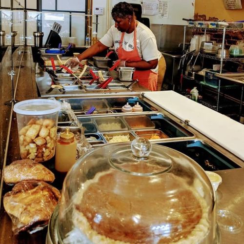 A woman prepares food in a busy kitchen, with various ingredients and dishes visible in the foreground and background.