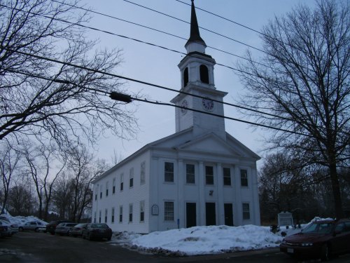 A white church with a tall steeple, surrounded by snow and parked cars, under a cloudy sky.