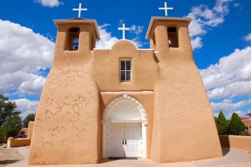 Adobe church with two bell towers and crosses, set against a blue sky with fluffy clouds.