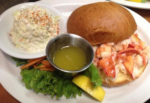 A lobster roll on a bun with a side of coleslaw, lettuce, lemon, and a small cup of melted butter.