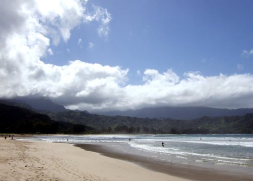 A serene beach scene with gentle waves, mountains in the background, and a partly cloudy sky.