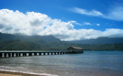 A serene beach scene with a wooden pier extending into calm waters, surrounded by lush green mountains and a blue sky.