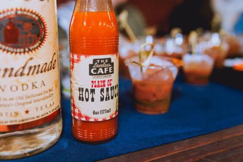 A bottle of vodka and a bottle of hot sauce on a table, with small cups in the background.