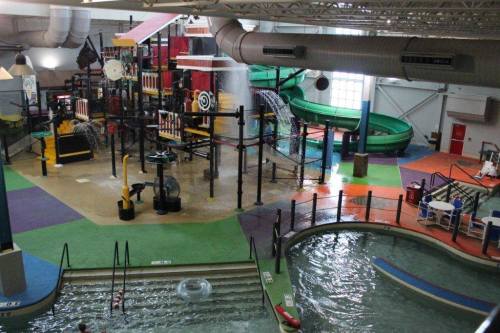 Indoor water park featuring colorful play structures, slides, and a lazy river area. Water splashes and kids play.
