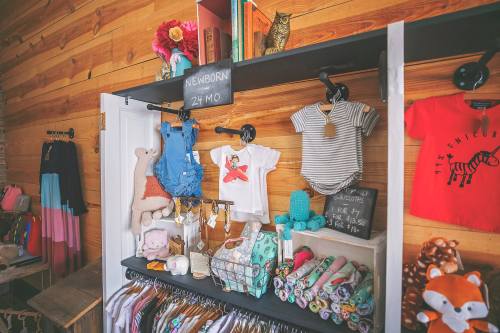 A display of children's clothing and accessories on shelves, featuring various outfits and plush toys against a wooden backdrop.
