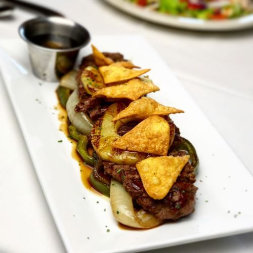 A platter of grilled beef with peppers and onions, topped with crispy tortilla chips and served with a small sauce cup.