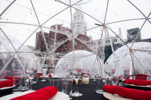 A cozy dining setup inside a transparent igloo, with festive lights and a snowy cityscape in the background.