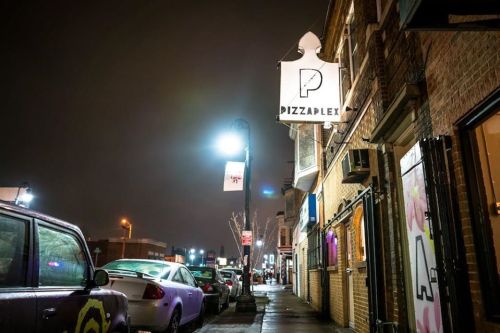A nighttime street view of Pizzaplex, with illuminated signs and parked cars along the sidewalk.