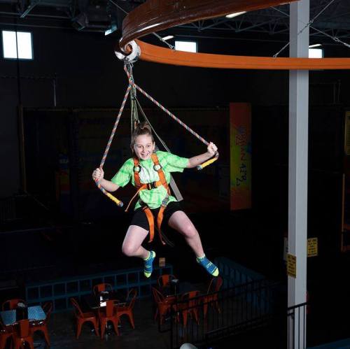 A girl in a green shirt is suspended in mid-air on a zip line at an indoor trampoline park.
