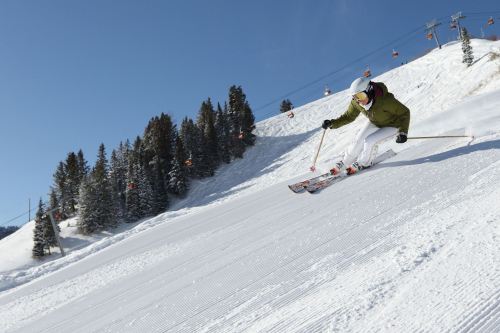 A skier in a green jacket glides down a snowy slope under a clear blue sky, with ski lifts in the background.