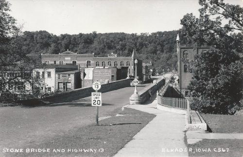 Black and white photo of Stone Bridge and Highway 13 in Elkader, Iowa, featuring buildings and a speed limit sign.