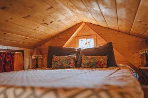 Cozy attic bedroom with a wooden ceiling, two decorative pillows, and a small window letting in natural light.