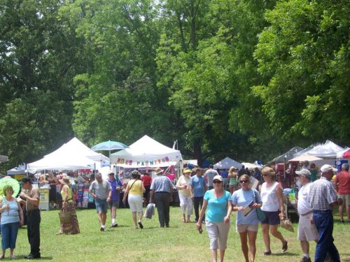 A bustling outdoor market with people browsing stalls under trees and tents on a sunny day.