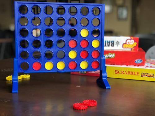 A blue Connect Four game with yellow and red discs, set on a table with various board games in the background.