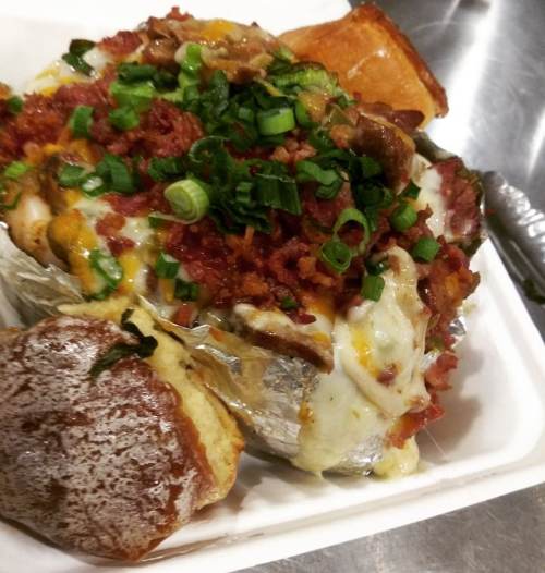 A loaded baked potato topped with cheese, bacon, green onions, and served with a soft roll on the side.