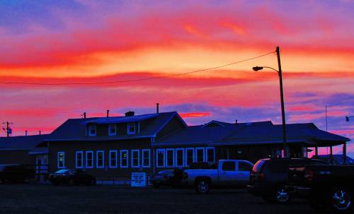 A colorful sunset casts vibrant hues over a building and parked cars, creating a serene evening scene.