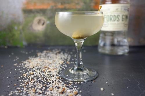 A cocktail in a coupe glass with an olive, surrounded by scattered grains and a bottle of Fords Gin in the background.