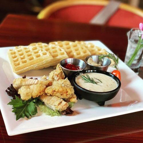 A plate featuring crispy fried chicken, waffles, and dipping sauces, garnished with lettuce and a cherry tomato.