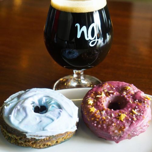 A glass of dark beer next to two colorful donuts on a plate, one with blue icing and the other with pink and sprinkles.