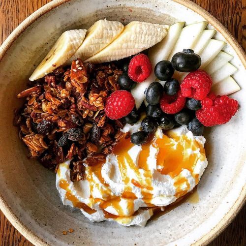 A bowl of yogurt topped with granola, banana slices, apple slices, raspberries, blueberries, and a drizzle of honey.