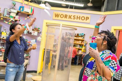 Two women in a colorful workshop space, one tossing an object into a clear container while the other watches excitedly.