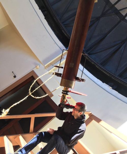 A person sits on stairs, holding a large telescope inside an observatory with a domed ceiling.