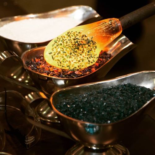A close-up of three metal bowls filled with colorful glass shards and a glowing glass tool resting on one.