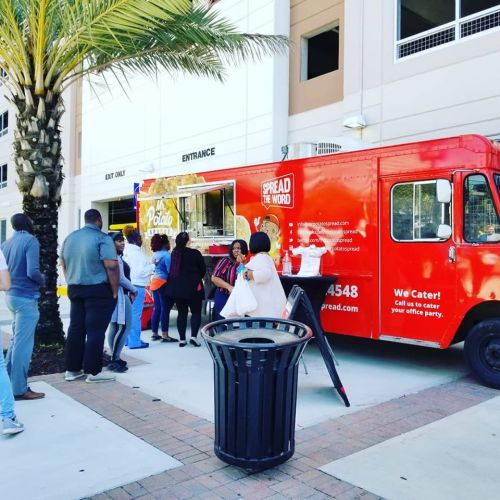 A red food truck with "Spread the Word" signage, serving customers lined up outside in a sunny area.