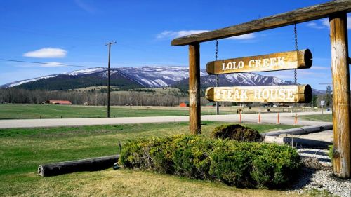 Sign for Lolo Creek Steak House with mountains in the background and a clear blue sky. Green grass in the foreground.