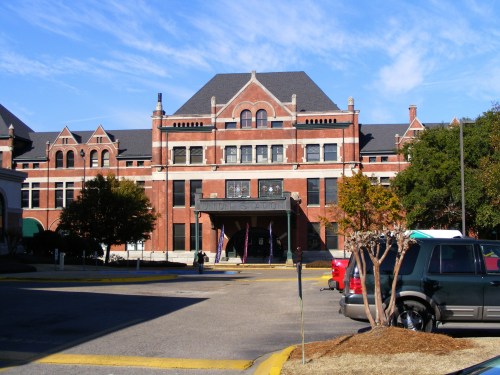 Historic brick building with a large entrance, surrounded by trees and parked cars under a clear blue sky.