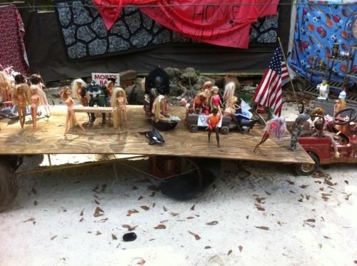A wooden platform displays various Barbie dolls in playful poses, surrounded by props and decorations in a whimsical setting.