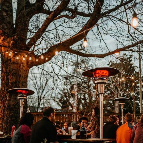 Outdoor dining scene with people gathered around tables, string lights, and patio heaters under a large tree.