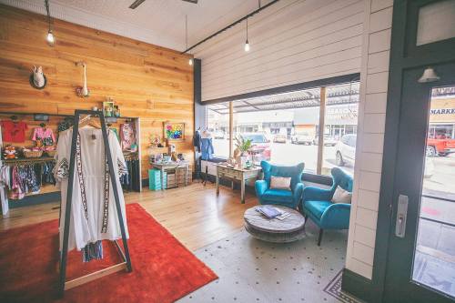 A cozy boutique interior featuring wooden walls, colorful clothing displays, and blue chairs by a window.