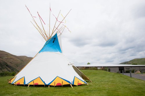 A colorful teepee with decorative patterns stands on green grass, set against a backdrop of rolling hills and cloudy skies.