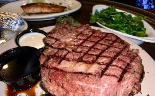 A juicy grilled steak with a crosshatch pattern, served with dipping sauces and sides of greens and fish.