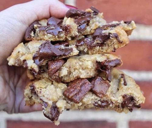 A hand holding stacked chocolate chip cookies with gooey chocolate chunks against a brick background.