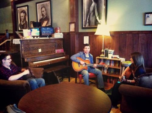 Three people in a cozy café setting, two playing guitars while one relaxes on a couch, with a piano in the background.