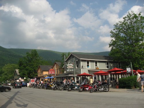 A quaint street scene with a restaurant, outdoor seating, and parked motorcycles, set against a backdrop of green mountains.