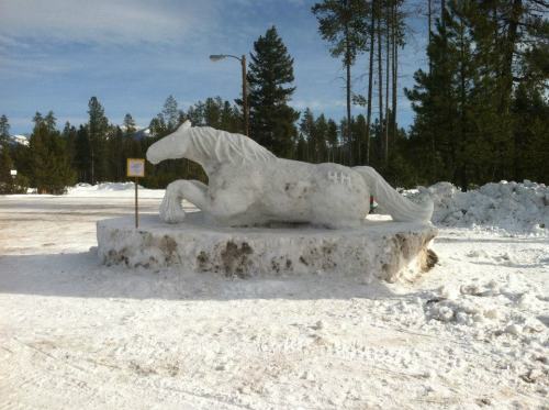 A large snow sculpture of a horse, positioned on a snowy base, surrounded by trees and a clear blue sky.