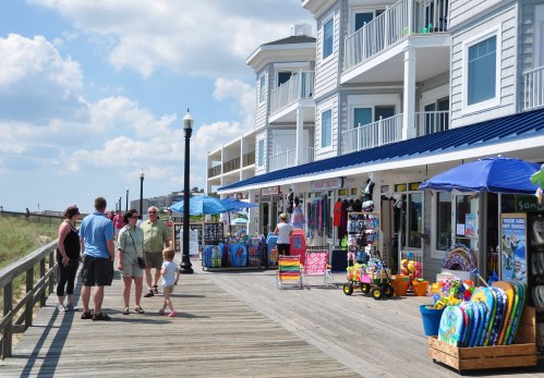 A boardwalk scene with people shopping at colorful beach shops and enjoying a sunny day by the coast.