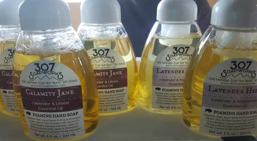 Bottles of foaming hand soap labeled "Calamity Jane" and "Lavender Hill," featuring essential oils, displayed on a table.