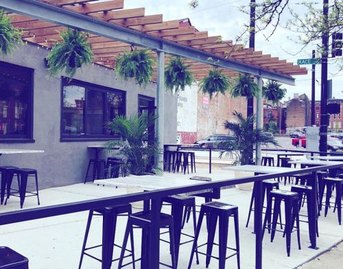 Outdoor dining area with black metal tables and stools, surrounded by hanging plants and a city street view.